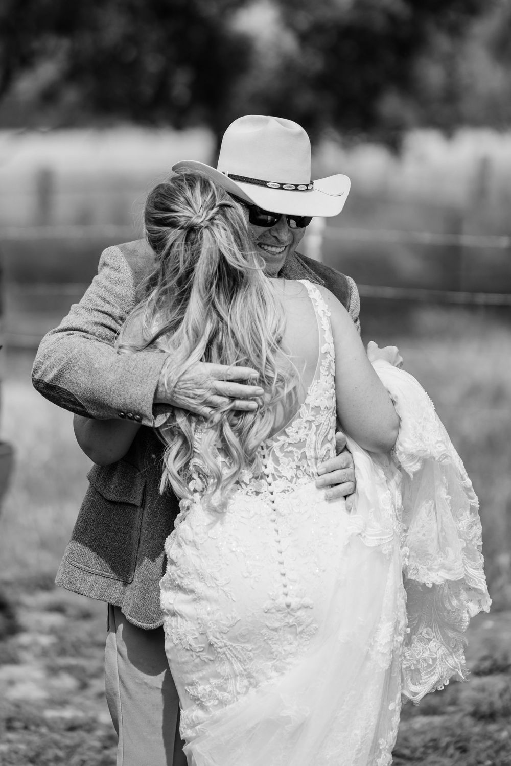 Black and white photo of a bride in lace gown embracing a man in a suit and cowboy hat at an outdoor wedding.