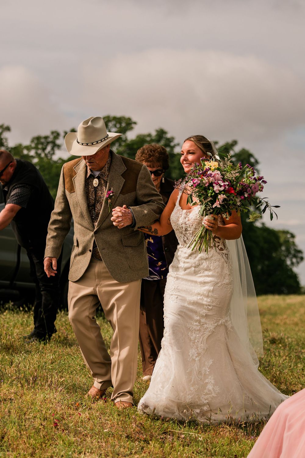 Bride in lace wedding dress walks down outdoor aisle holding colorful bouquet, escorted by man in cowboy hat and tan suit.