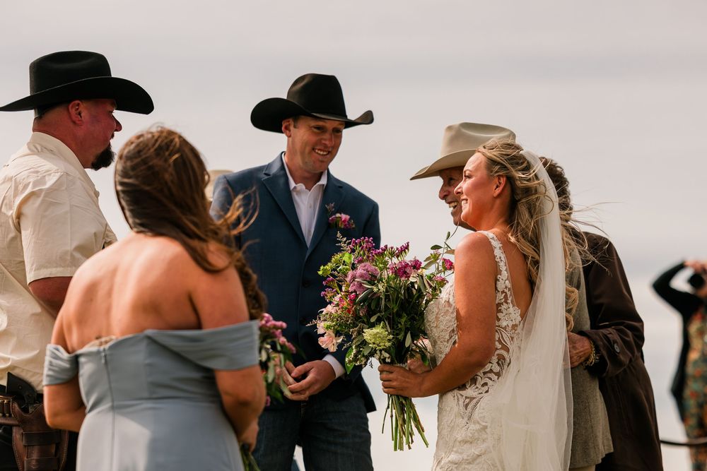 Outdoor western wedding ceremony with bride holding flowers, groom in blue suit, and guests in cowboy hats.