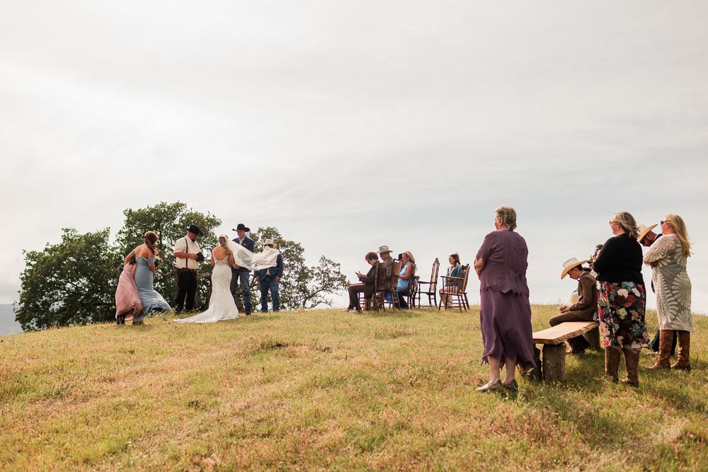 Outdoor hilltop wedding ceremony with guests gathered around couple exchanging vows under overcast sky.