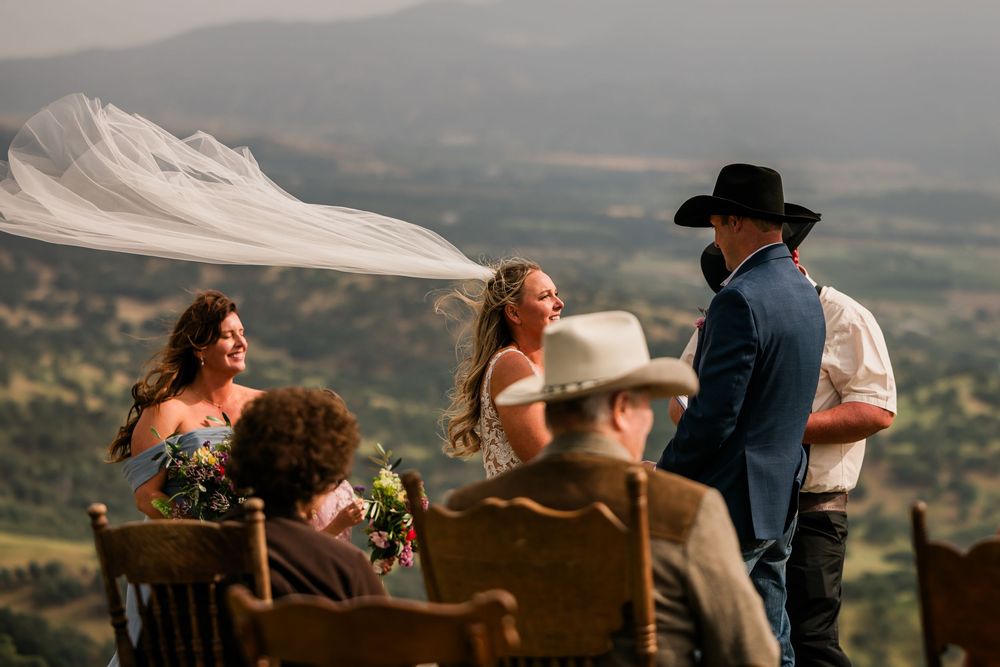 Outdoor wedding ceremony on a hilltop with guests seated, bride's veil blowing in wind, scenic mountain valley backdrop.