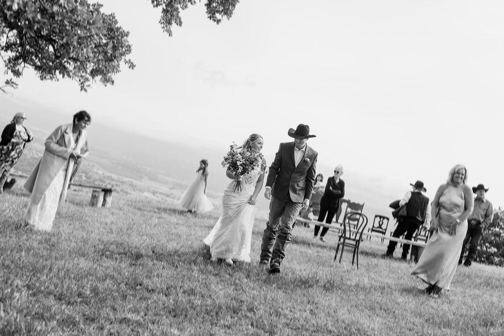 Black and white outdoor wedding ceremony on a grassy hill with guests, bride in white gown, groom in cowboy hat.
