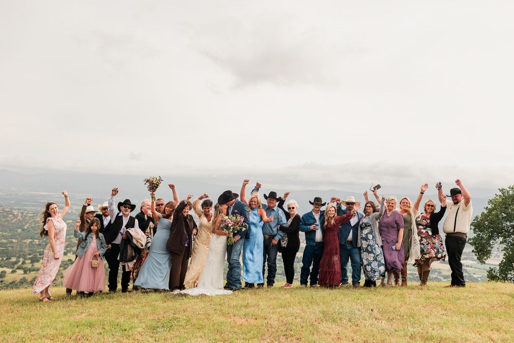 Wedding party celebrating outdoors on a hilltop with scenic views, raising arms in joy on a cloudy day.