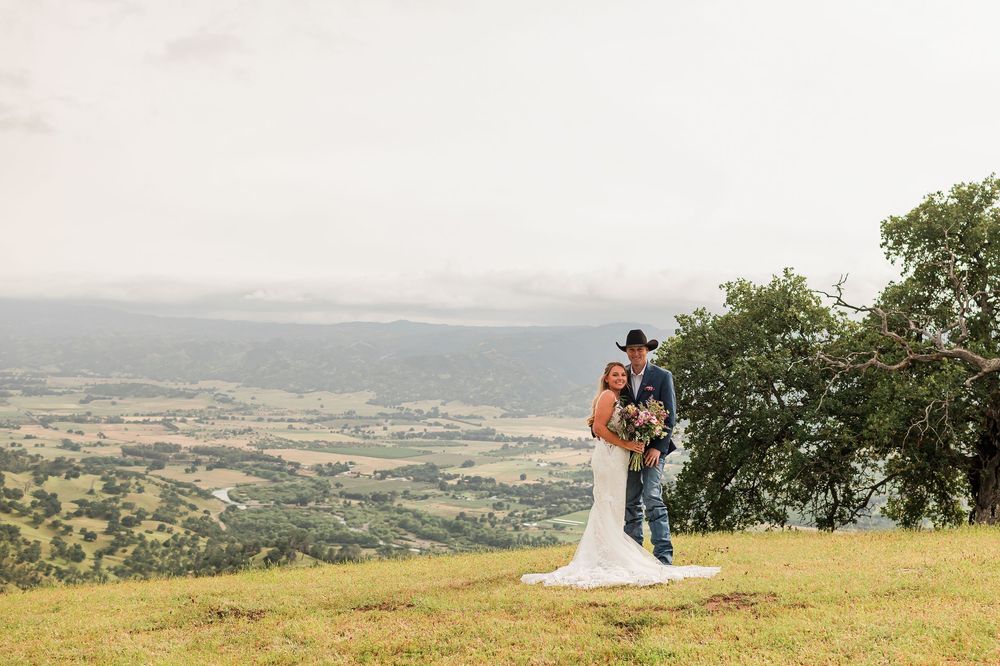 Bride and groom embrace on hilltop with stunning valley views, groom wearing cowboy hat at outdoor wedding.