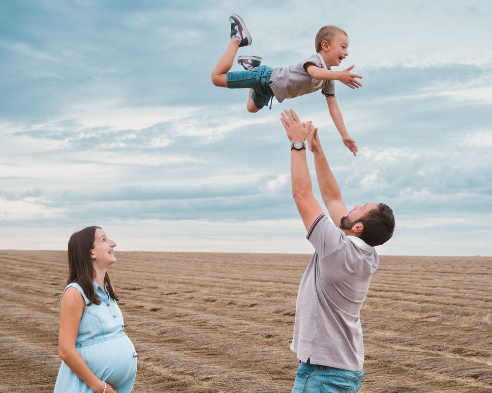 Père soulevant un jeune enfant dans les airs, mère enceinte à côté, champ de lin coupé