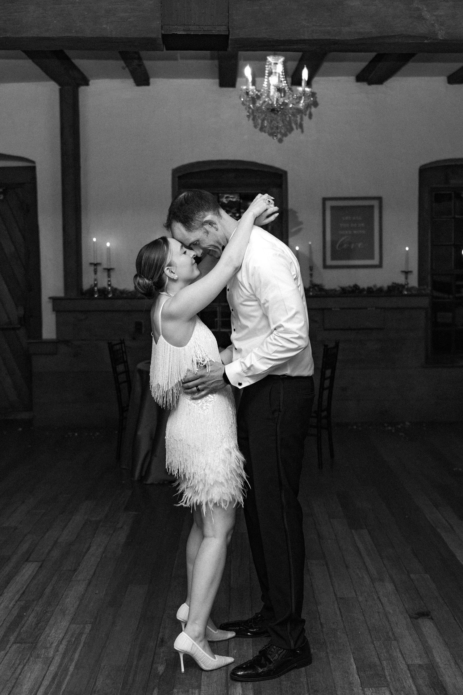 Black and white photo of bride and groom sharing intimate slow dance under chandelier at reception.