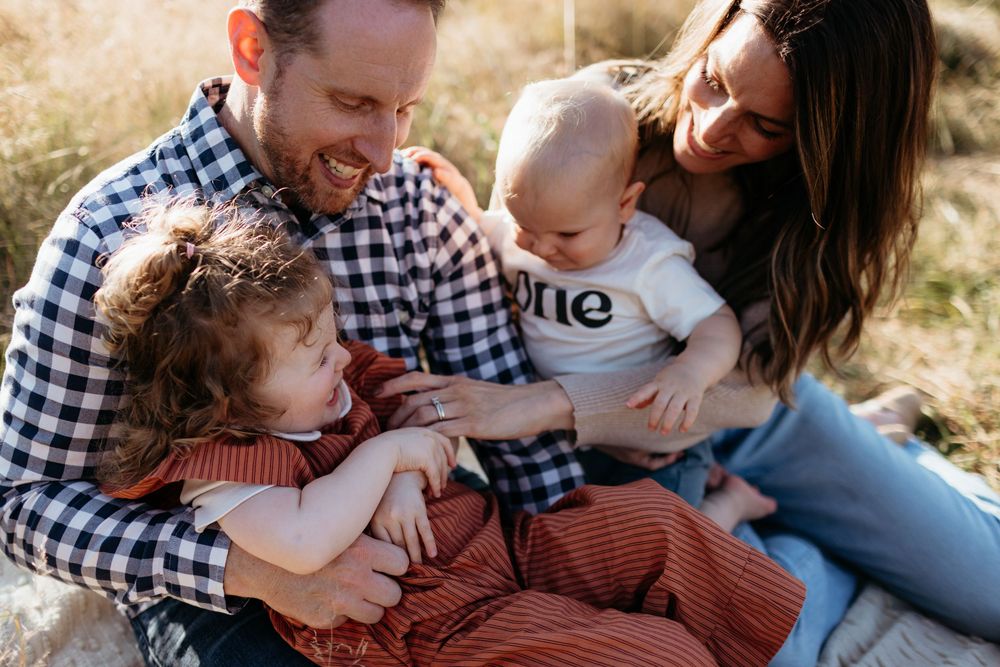 Happy family of four sitting outdoors, parents smiling at baby wearing 'one' onesie with toddler in red dress.