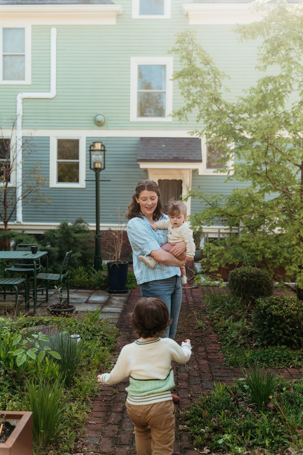 A mother holding a baby smiles at a toddler in a lush garden outside a charming blue house.