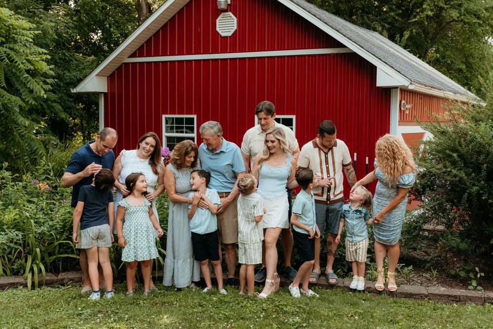 Large family group smiling together outdoors in front of a red barn surrounded by lush green trees.