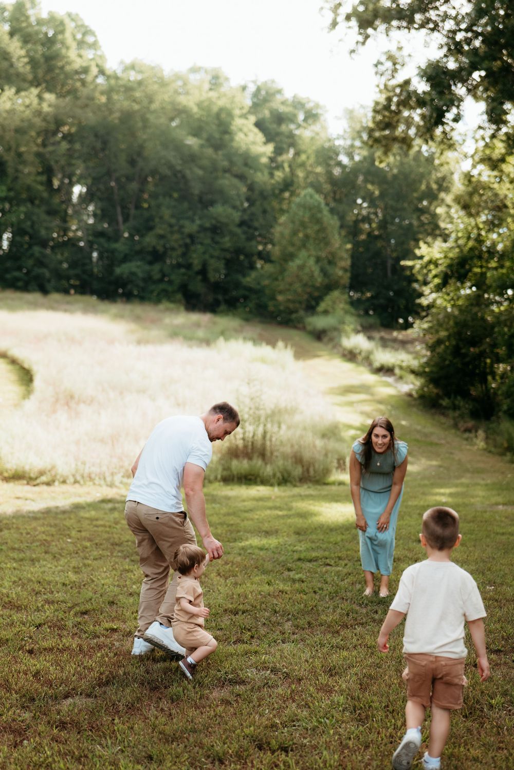 Family enjoying outdoor playtime in a sunny green meadow, parents helping toddler walk while older child plays nearby.