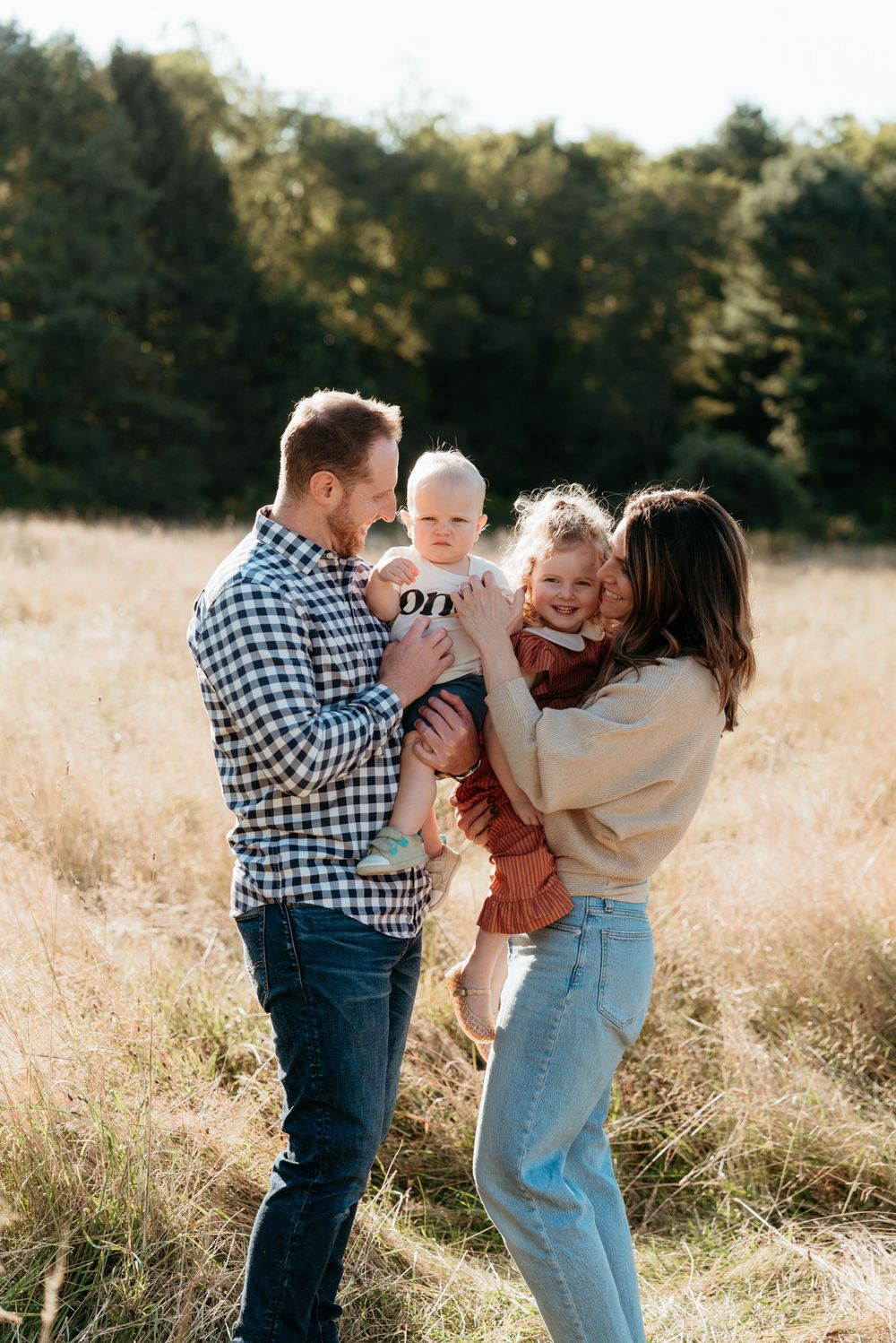 A happy family of four poses in a sunny autumn field, parents holding two young children while smiling outdoors.