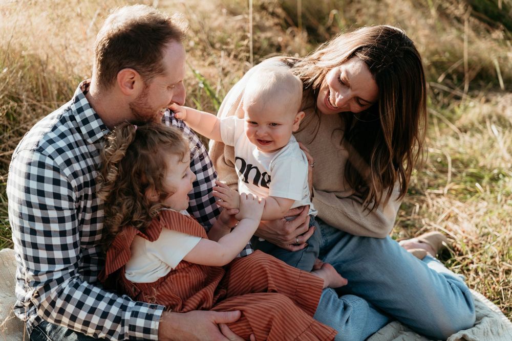 Happy family of four sitting outdoors in golden grass, parents smiling at baby and toddler girl during fall photo session.