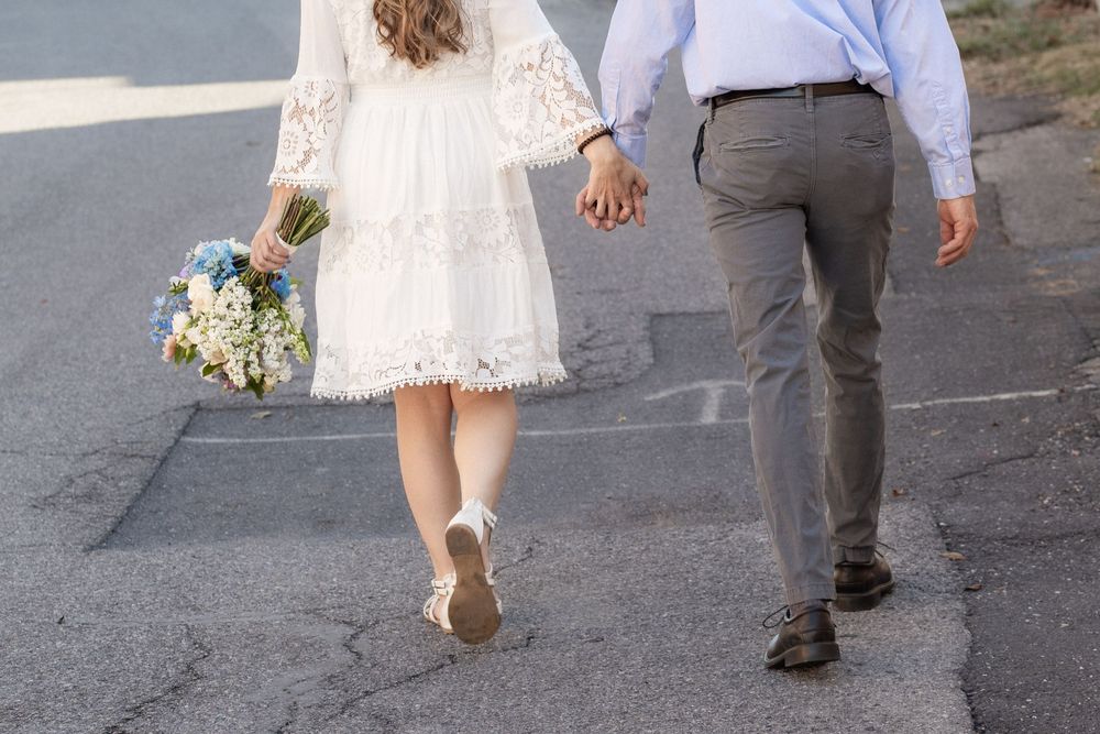 Bride in white lace dress holding blue bouquet walks hand-in-hand with groom in gray pants on pavement.