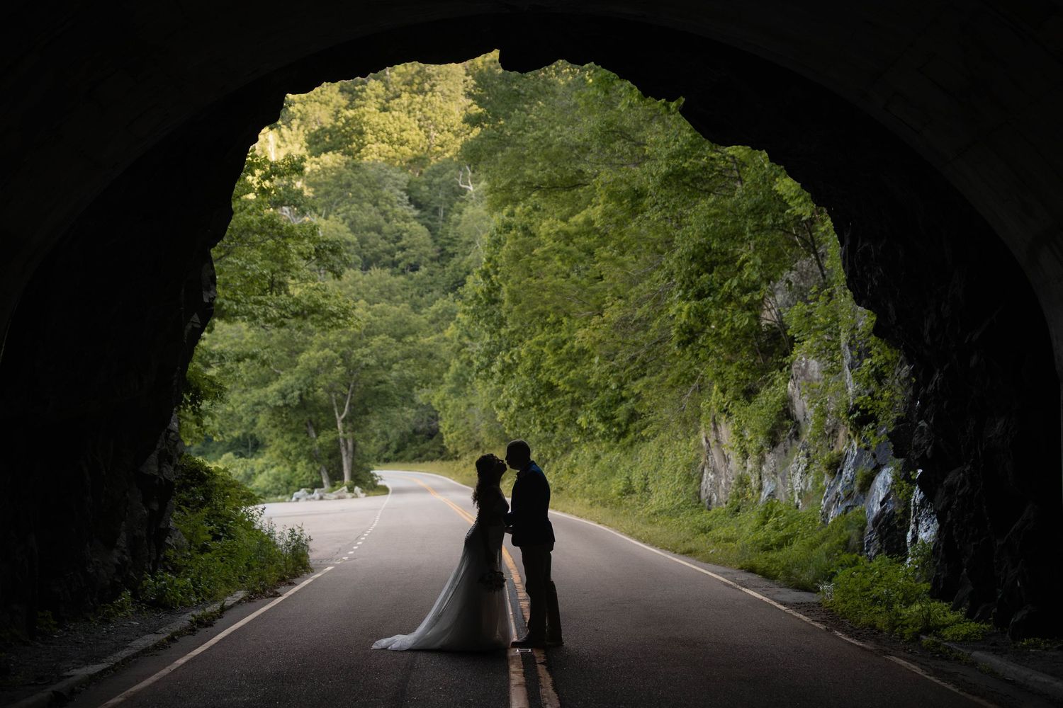 Bride and groom silhouetted in a tunnel entrance framed by lush green trees on a scenic road.