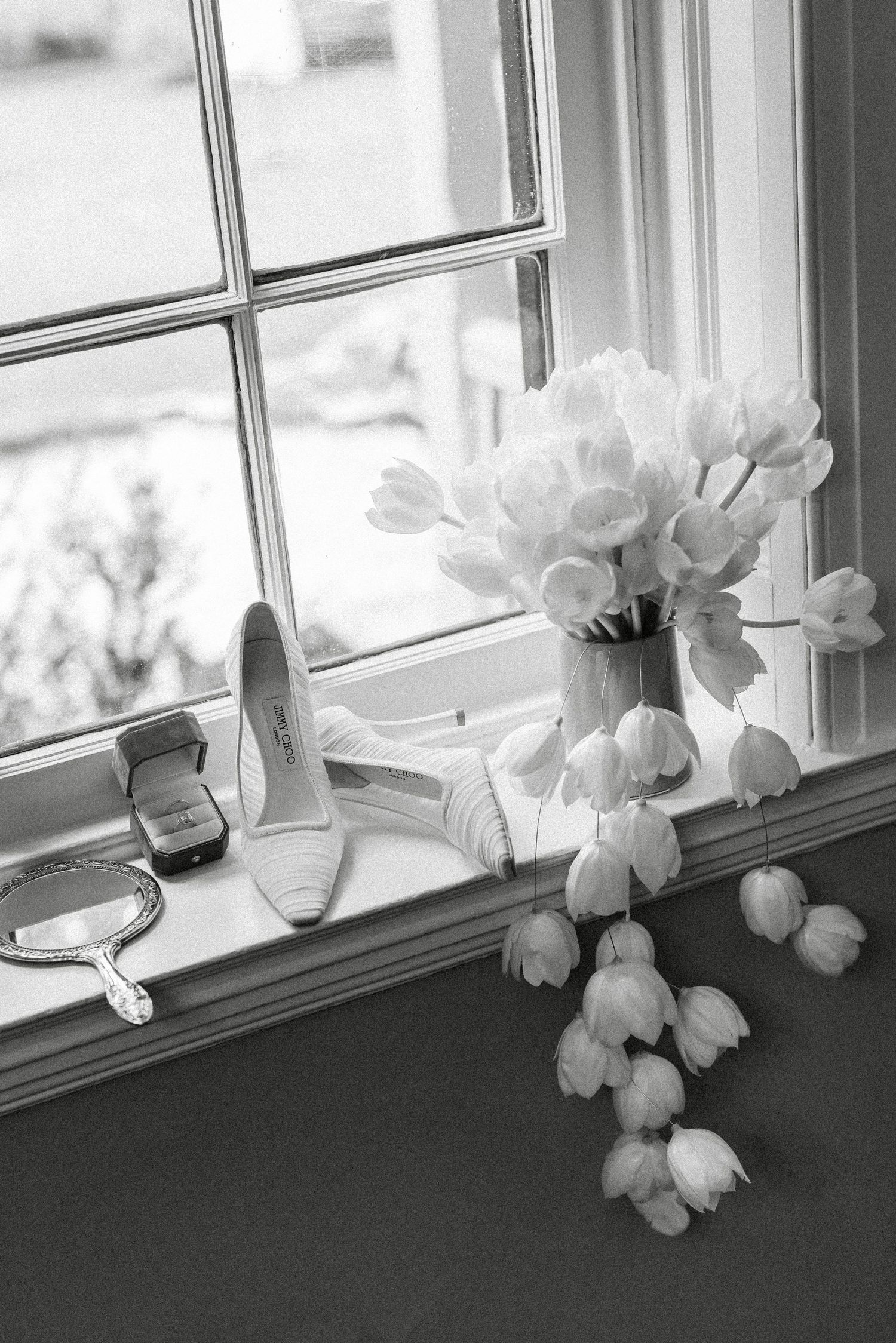 Black and white photo of bridal accessories including heels, tulip bouquet, and mirror on a windowsill.