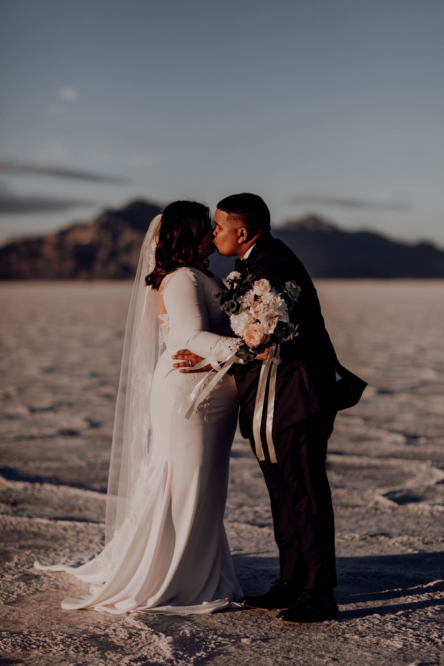 Couple sharing a kiss in wedding attire on the Bonneville Salt Flats with mountains at sunset.