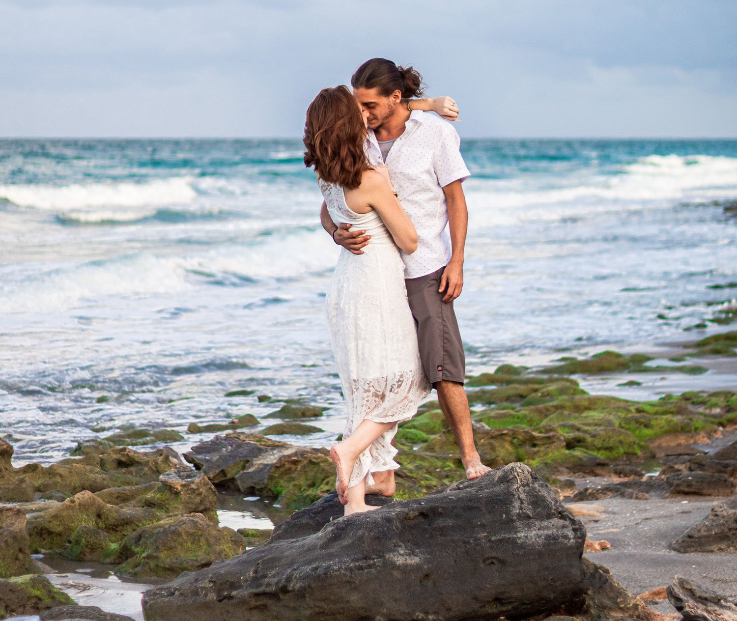 Couple in black attire standing on rocky shore at sunset, woman's flowing dress blowing in the wind with ocean waves behind at blowing rocks