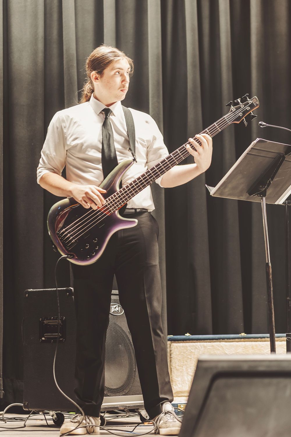 Young male musician playing a purple bass guitar on stage, wearing white shirt and black tie with music stand nearby.