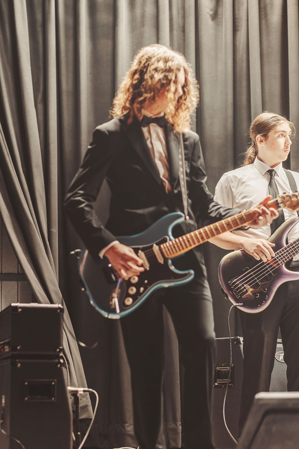 Two guitarists in formal attire perform on stage, one with curly hair in a black suit, the other in a white shirt and tie.