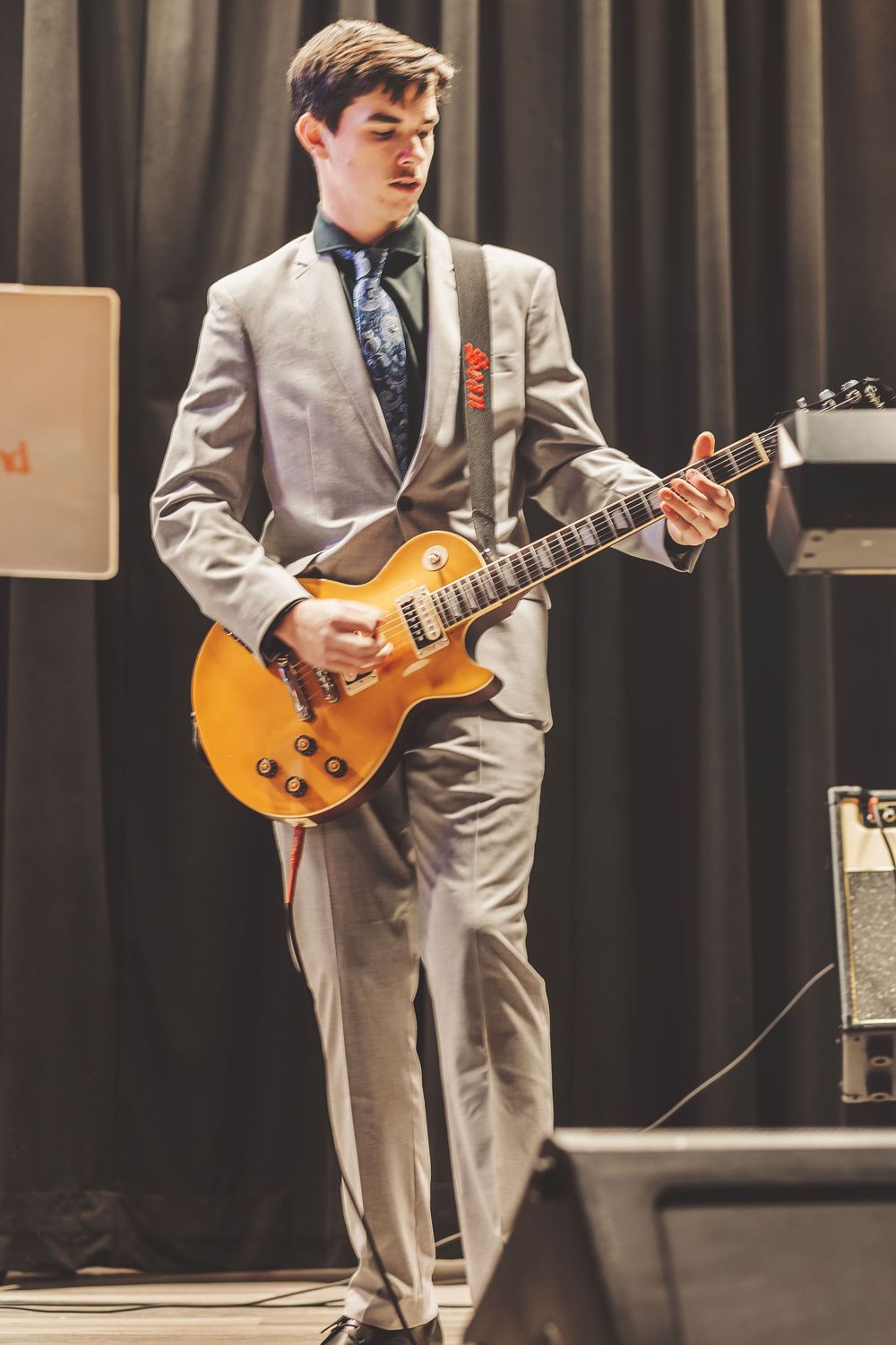 Young man in gray suit playing a gold Les Paul electric guitar on stage during a live performance.