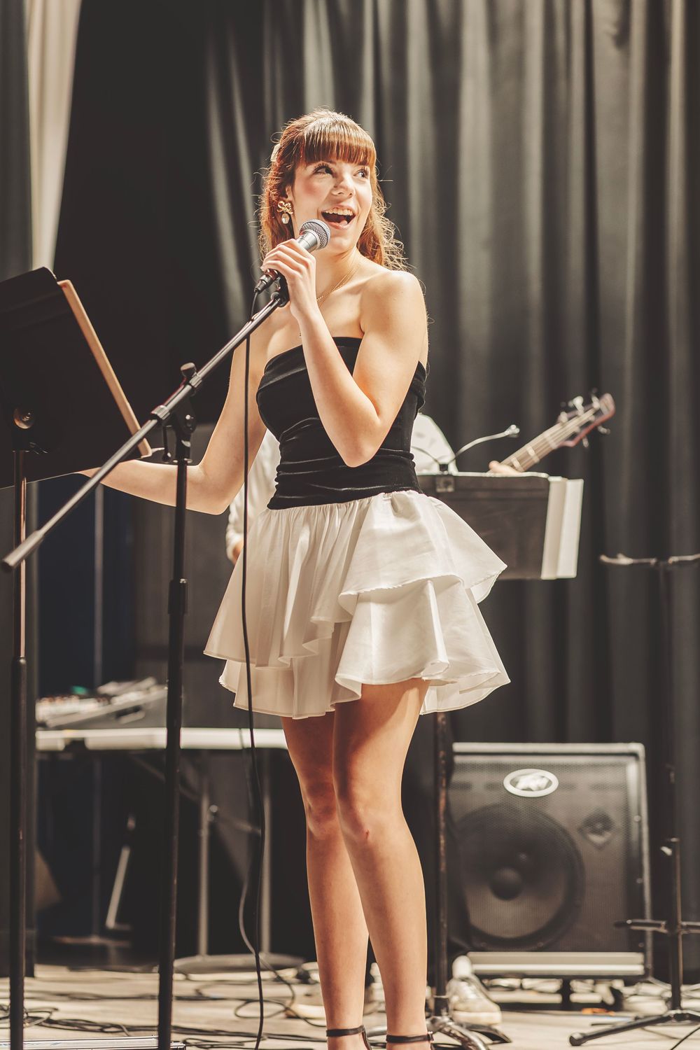 Young woman in black and white dress singing into microphone on stage with band equipment in background.