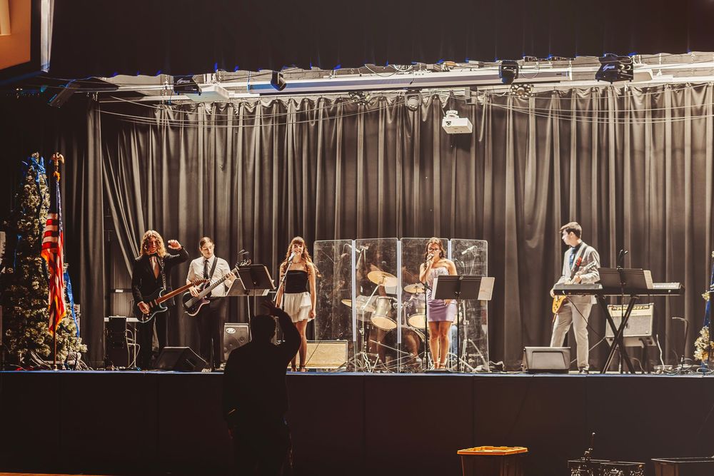 Live band performing on stage with guitars, keyboard, and saxophone at an indoor venue with holiday decorations.