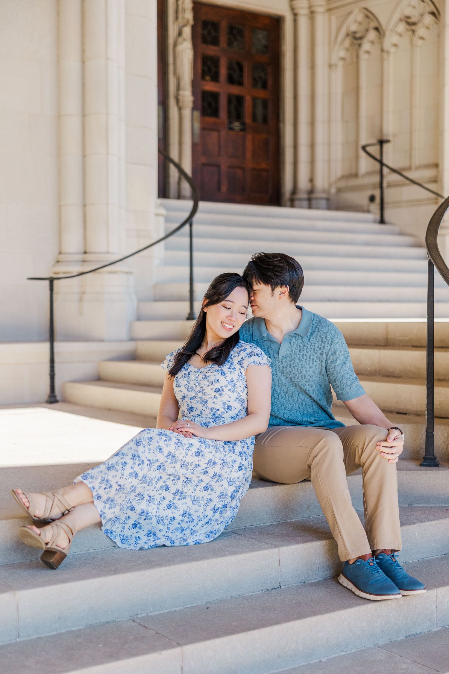 A couple sits on stone steps outside a historic building, the man whispering to the smiling woman in a blue floral dress.