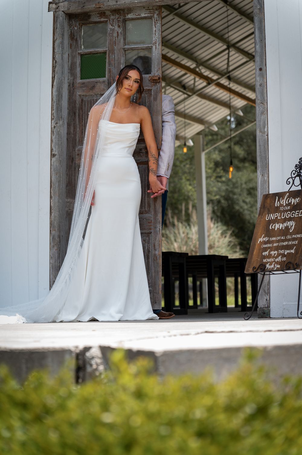 Bride in white gown and veil holds groom's hand at rustic wooden chapel doors beside an unplugged ceremony sign.