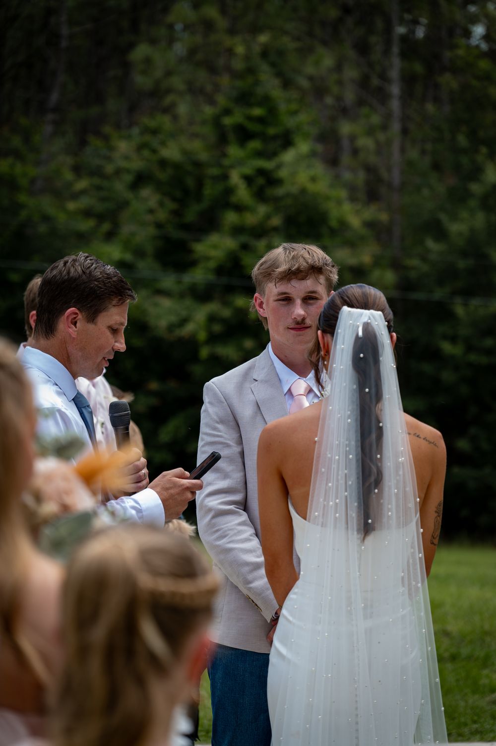 Young groom in gray suit exchanges vows with bride in white gown and veil at outdoor wedding ceremony.