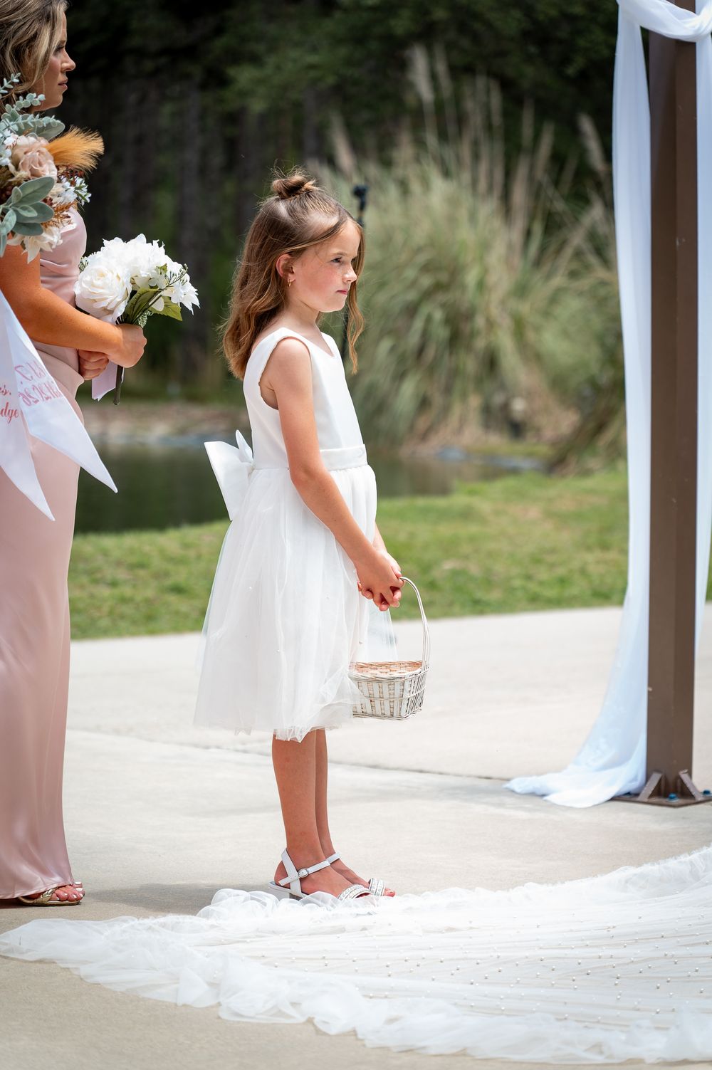 Young flower girl in white dress holding wicker basket stands at wedding ceremony altar.