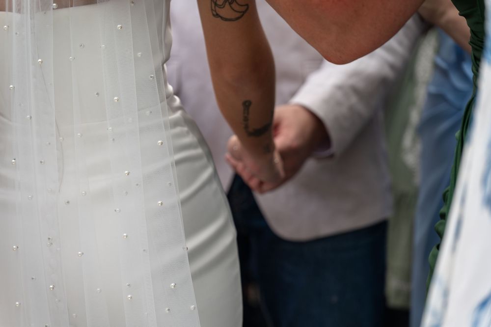Close-up of bride in white wedding dress standing next to person with cross tattoo on forearm.