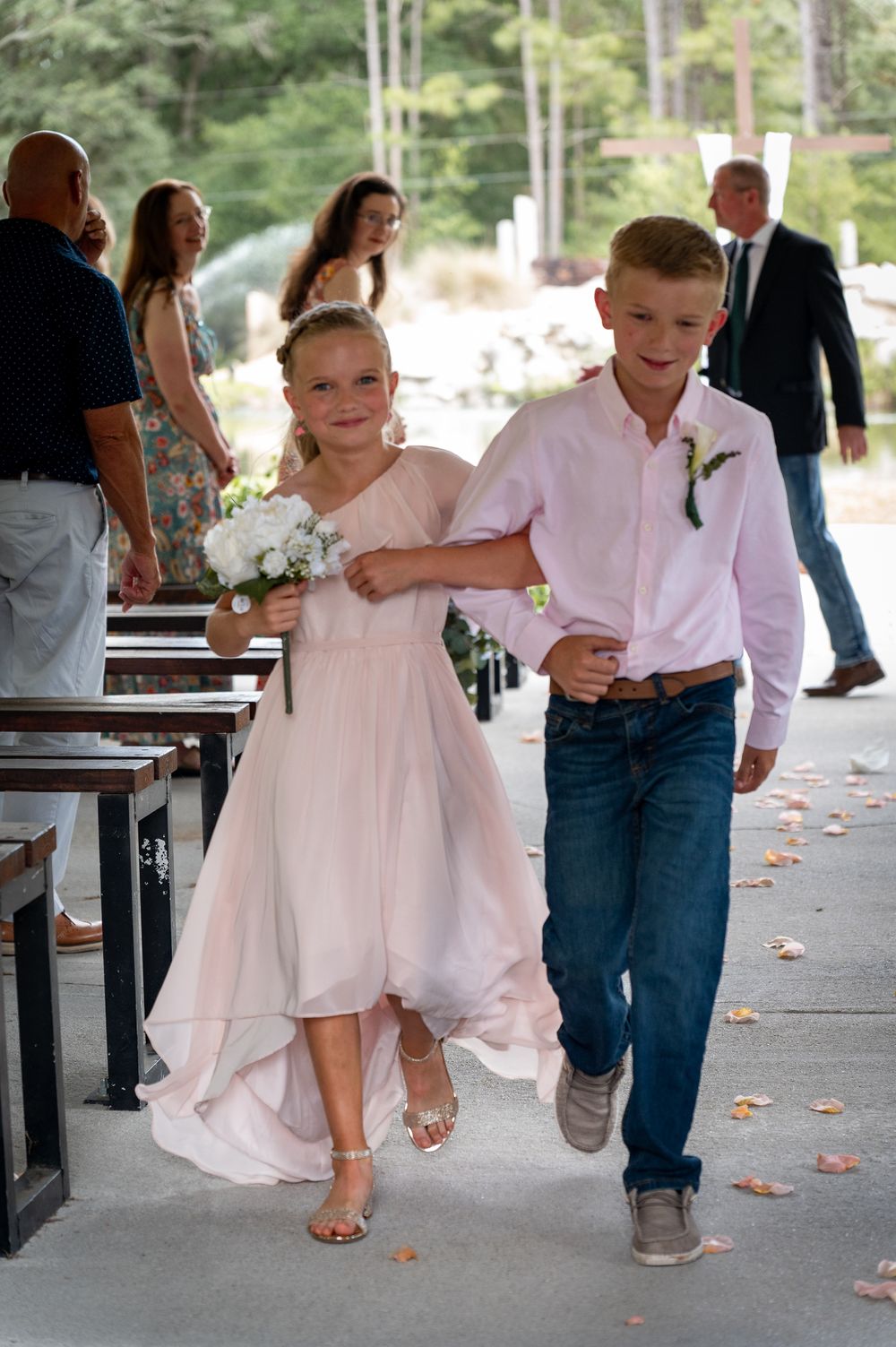 Young flower girl in pink dress and ring bearer in pink shirt walk down wedding aisle together.