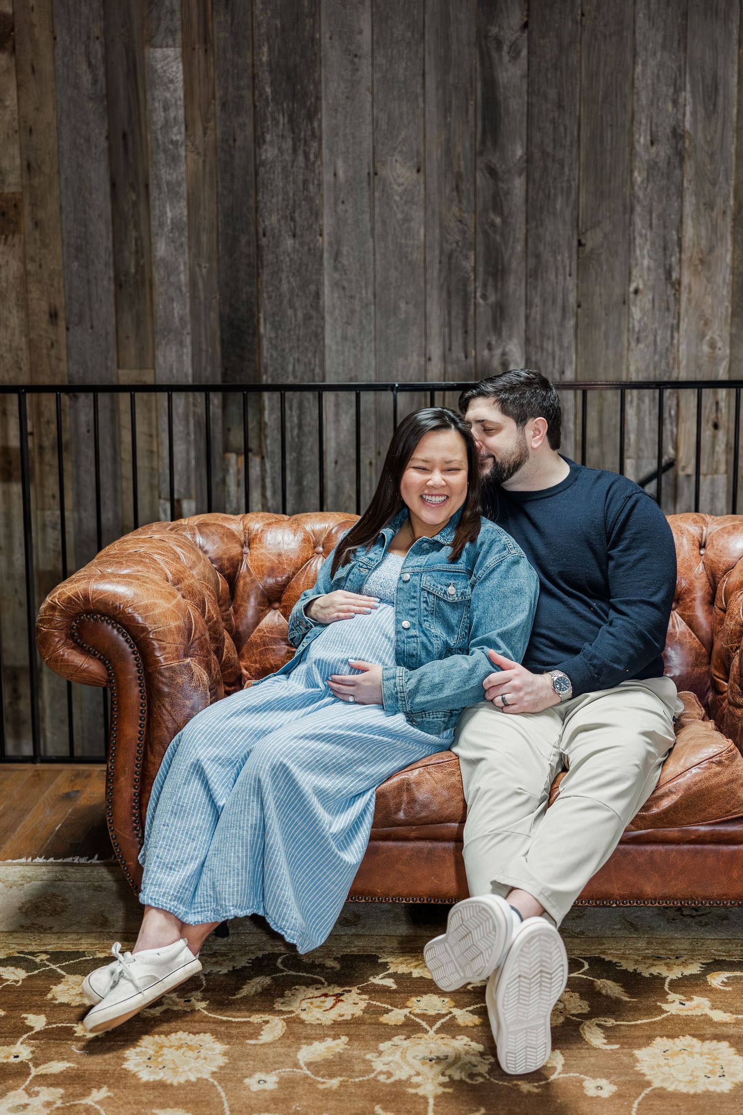 Pregnant woman in denim jacket sits with partner on leather couch, both smiling at her baby bump.