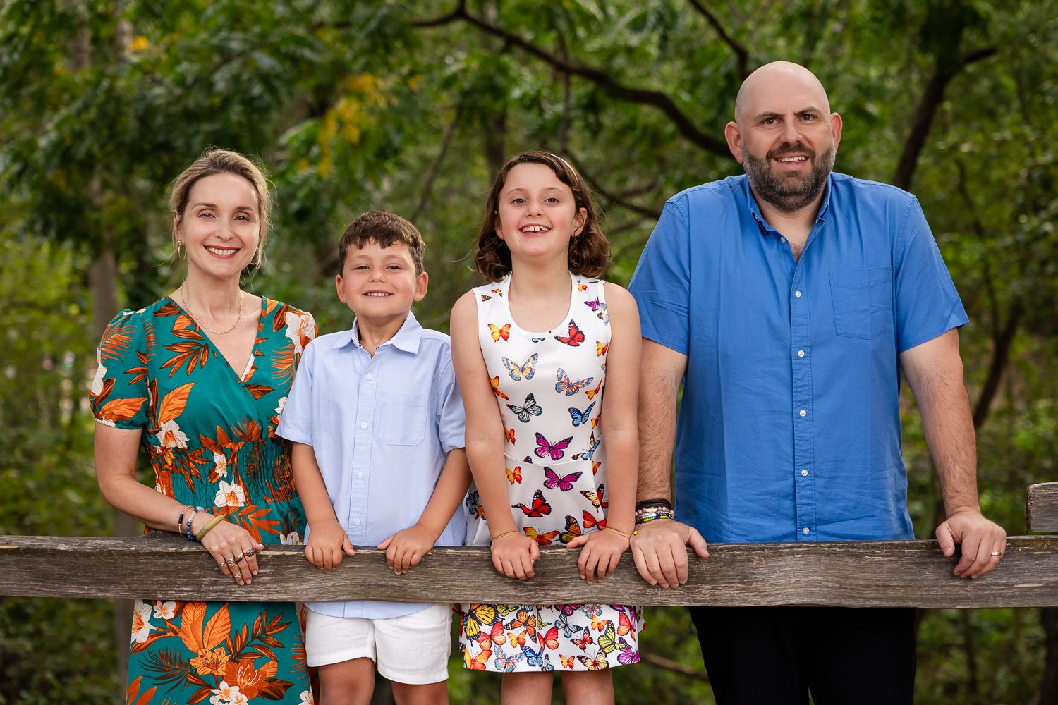 Happy family of four smiling together outdoors, posing by a wooden fence surrounded by lush green trees.
