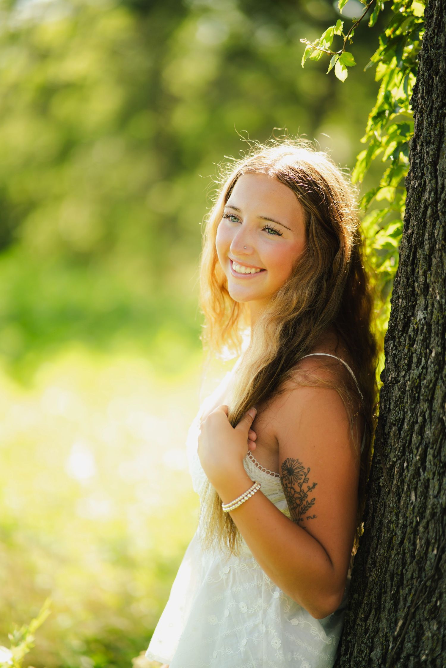 Smiling young woman with long hair and tattoo leans against tree in sunny green park wearing white dress.
