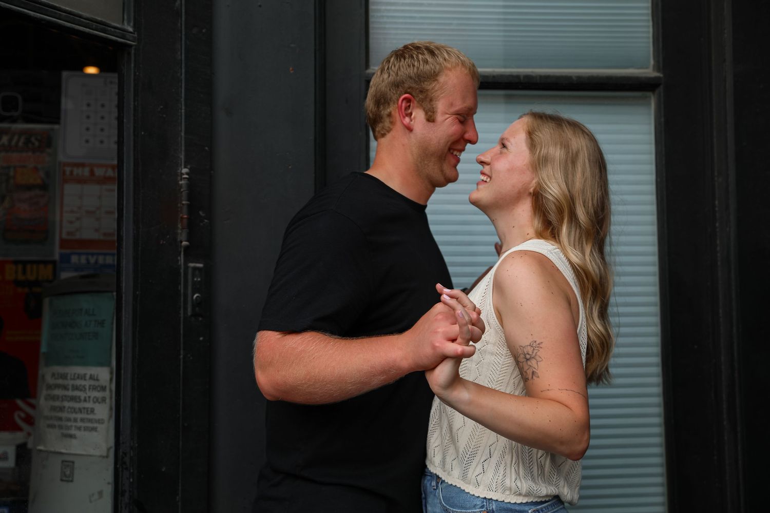 A smiling couple holds hands and gazes at each other outside a building on a sunny day.