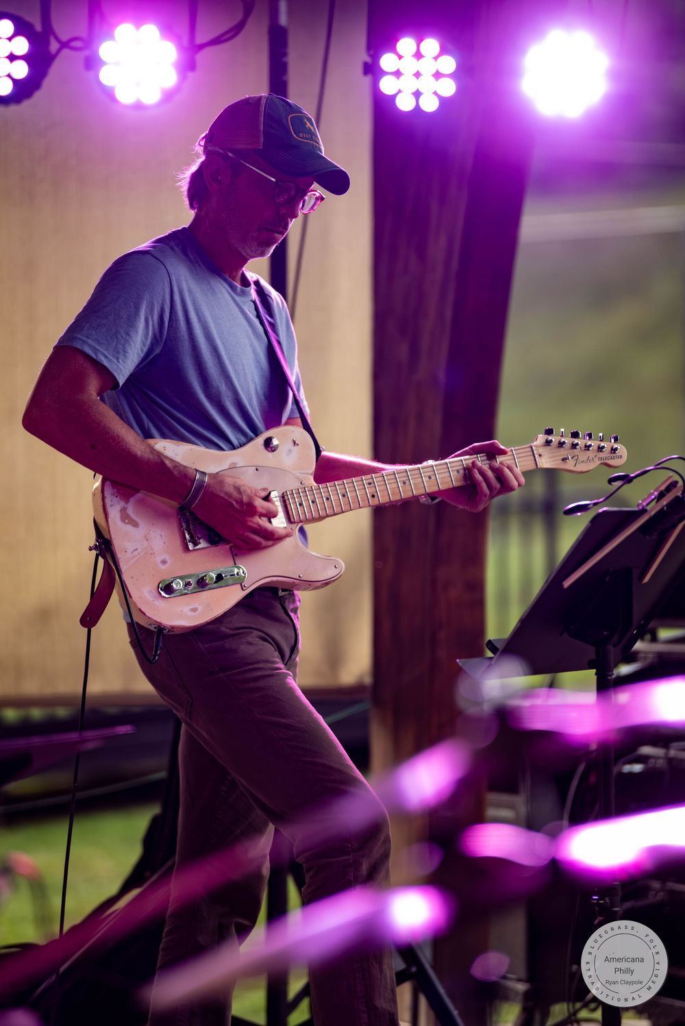 Guitarist performing on stage under purple lights, playing a pink electric guitar at an outdoor concert.