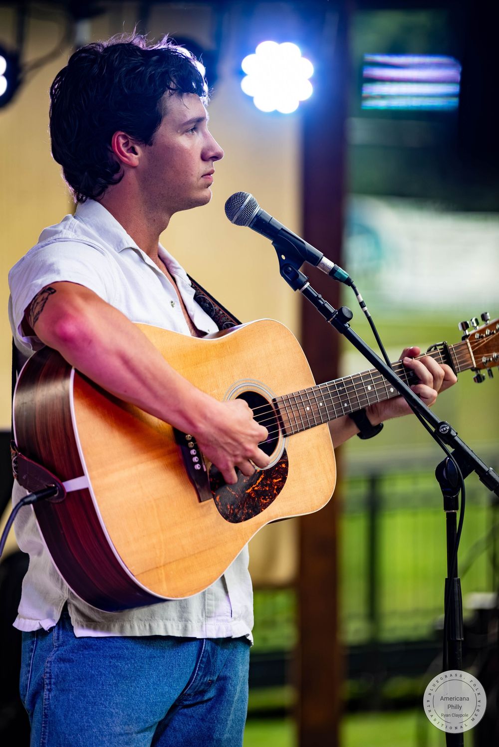 Young man playing acoustic guitar and singing into a microphone on stage under bright lights.