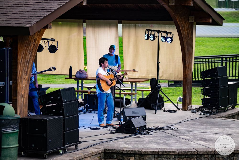 Musician playing acoustic guitar on an outdoor pavilion stage with speakers and lighting equipment setup.