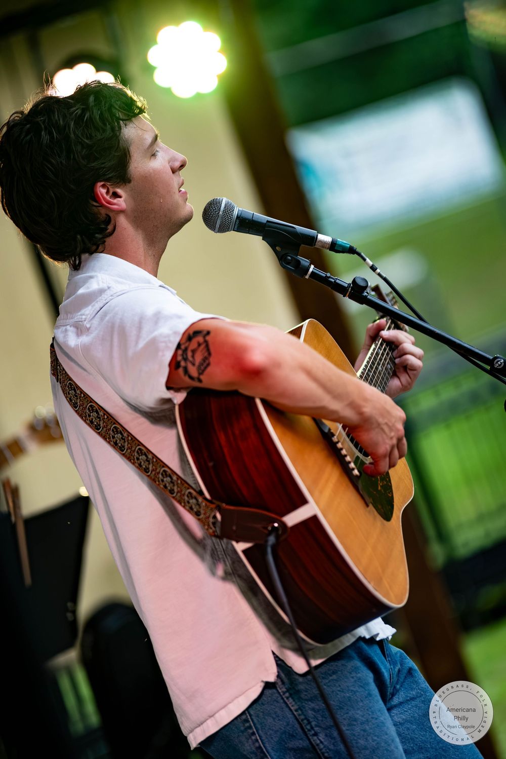 Young male musician plays acoustic guitar and sings into microphone, wearing white shirt with tattoo visible on arm.