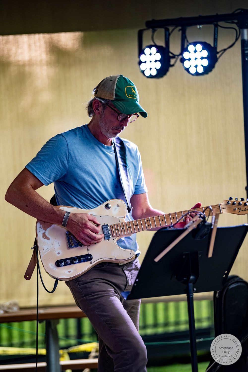 Man in green cap plays white electric guitar on outdoor stage with blue stage lights behind him.