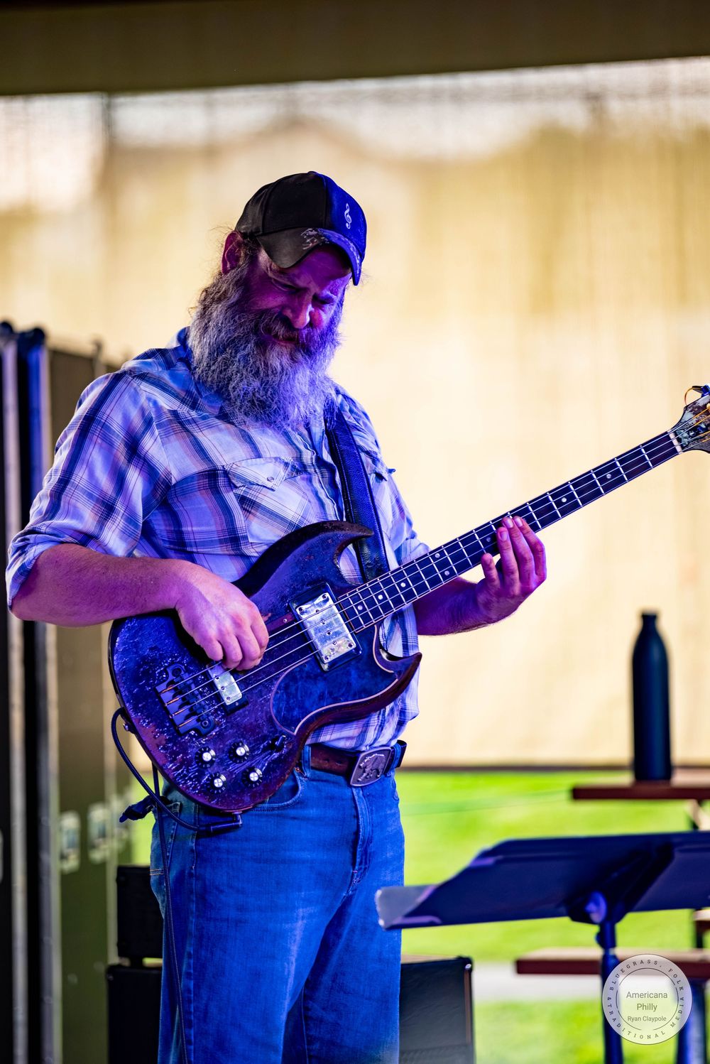 Bearded man playing electric bass guitar on stage wearing a plaid shirt and black cap at an outdoor concert.