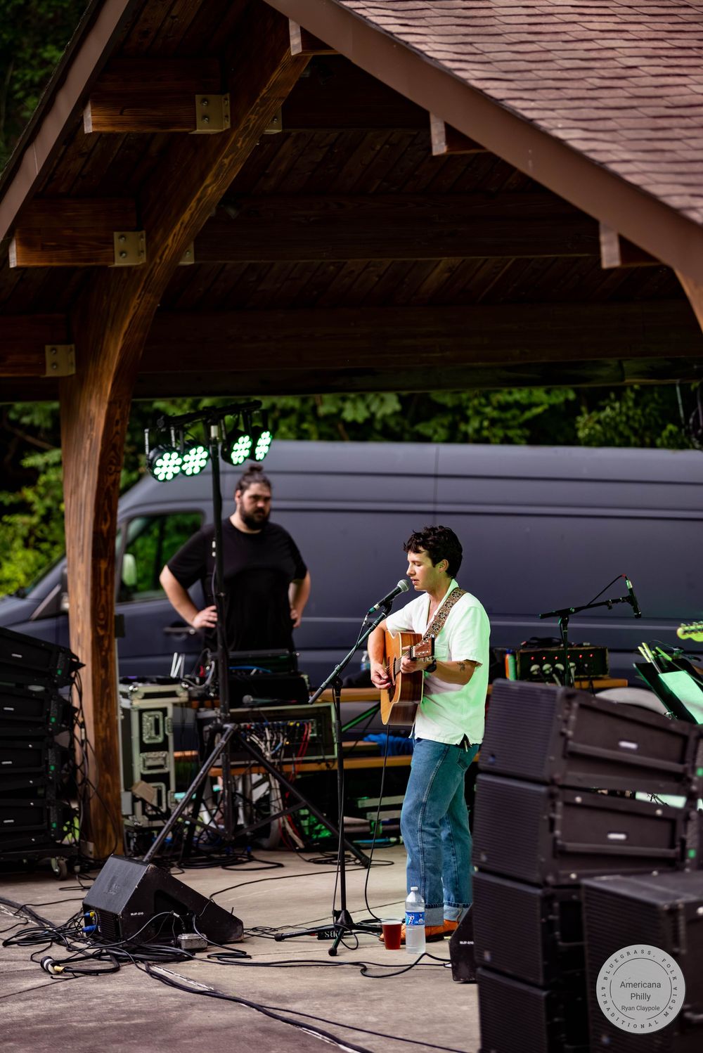 Musician playing acoustic guitar at outdoor stage microphone with sound technician standing behind at a park pavilion.