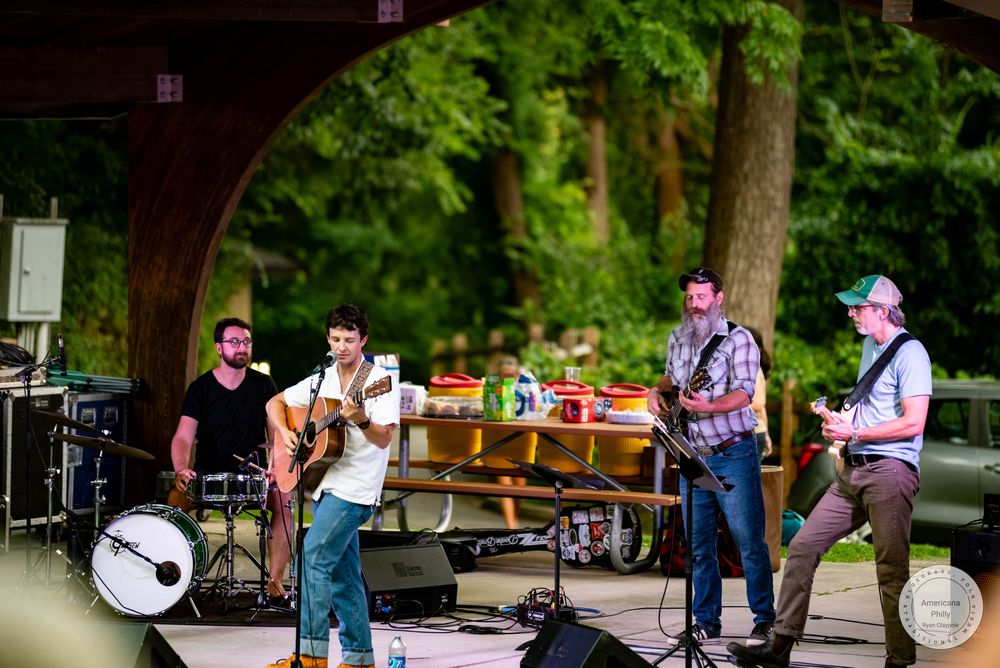 Four-piece band performs outdoor concert on wooded stage, playing guitars and drums at a summer music festival.