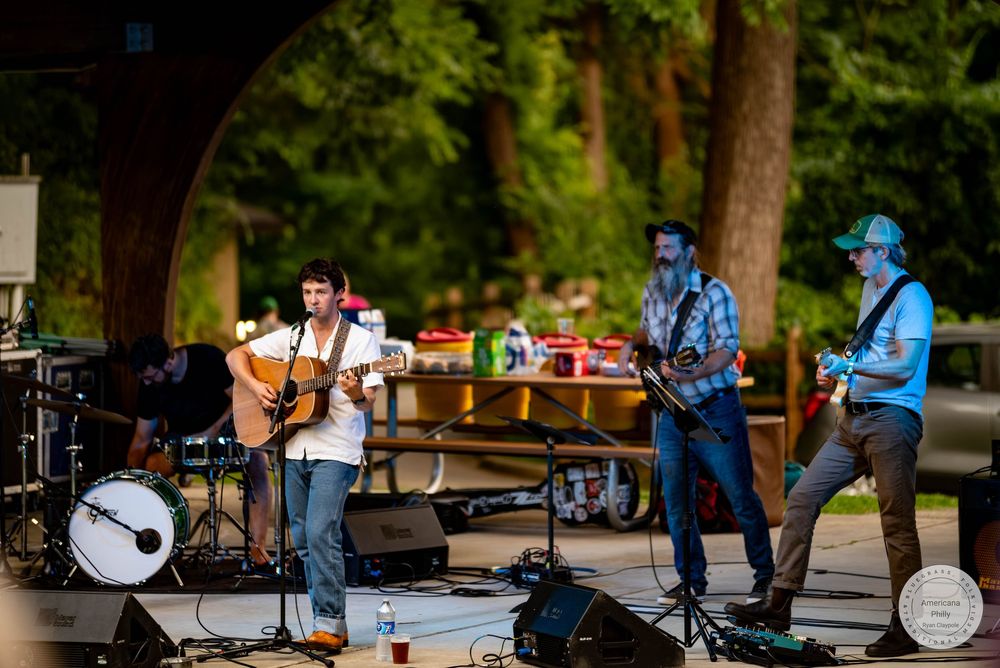 Three musicians perform outdoors on a shaded stage, playing guitars at a casual summer concert.