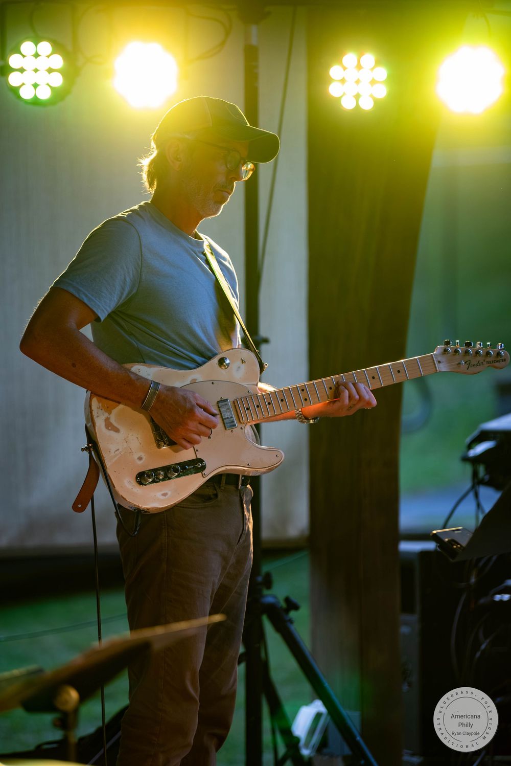 Guitarist performing on stage under green and yellow lights, wearing a cap and blue t-shirt, playing a white electric guitar.
