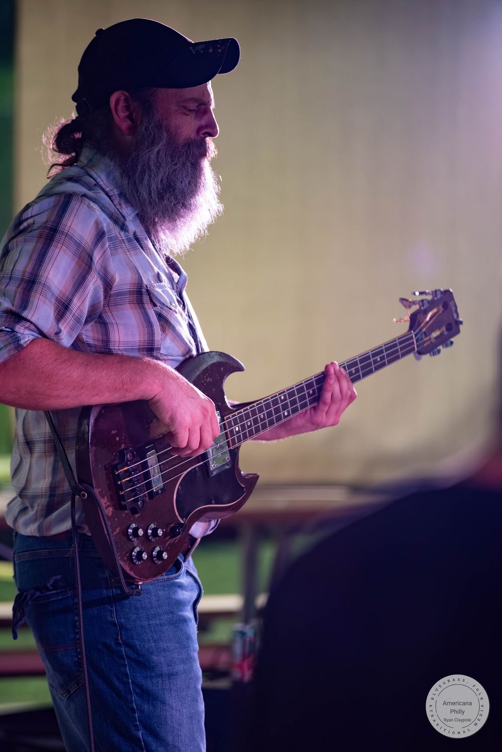 Bearded man playing electric bass guitar on stage wearing plaid shirt and black cap at live music event.