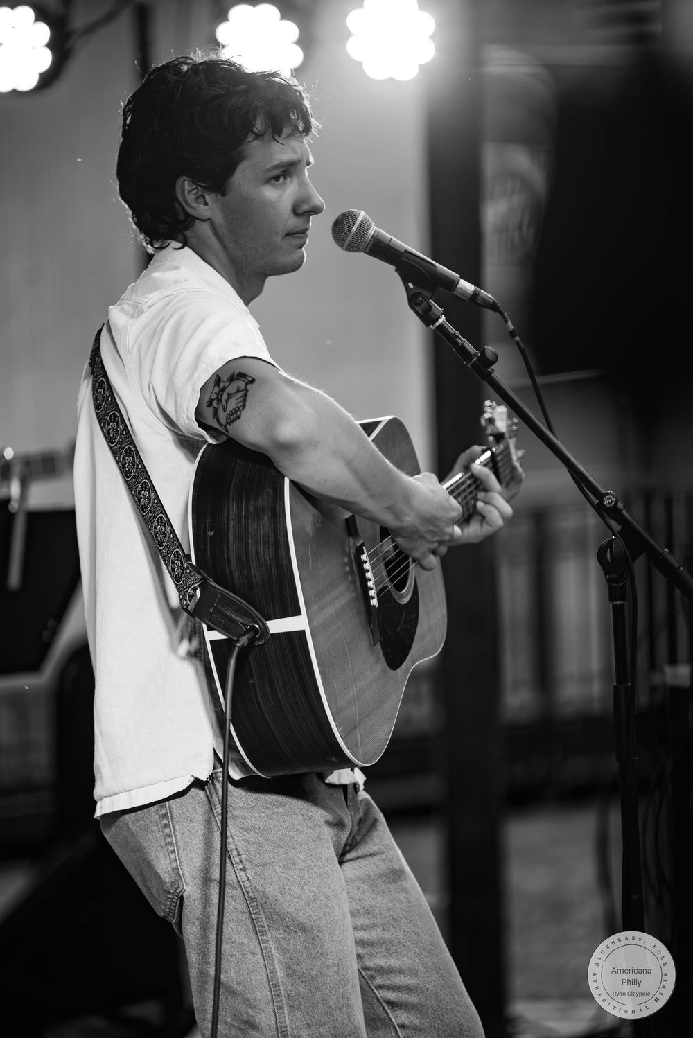 Young man playing acoustic guitar at microphone stand, black and white concert photo, tattoo visible on arm.