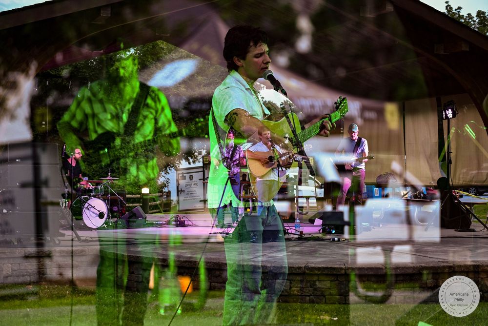 Live band performing on outdoor stage, guitarist in green shirt plays music at a park concert event.