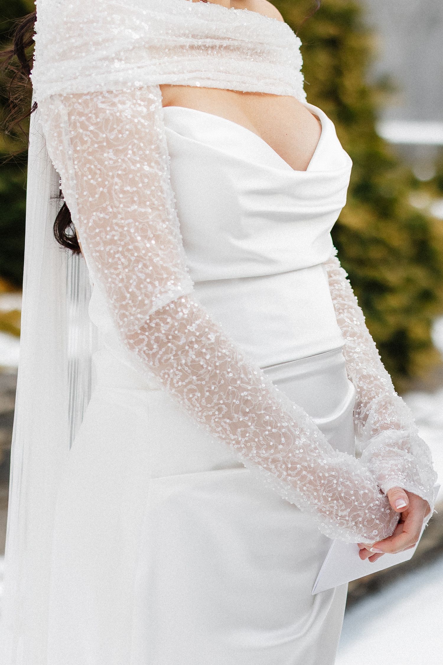 Bride in elegant white wedding dress with lace sleeves and veil, standing outdoors in winter setting.